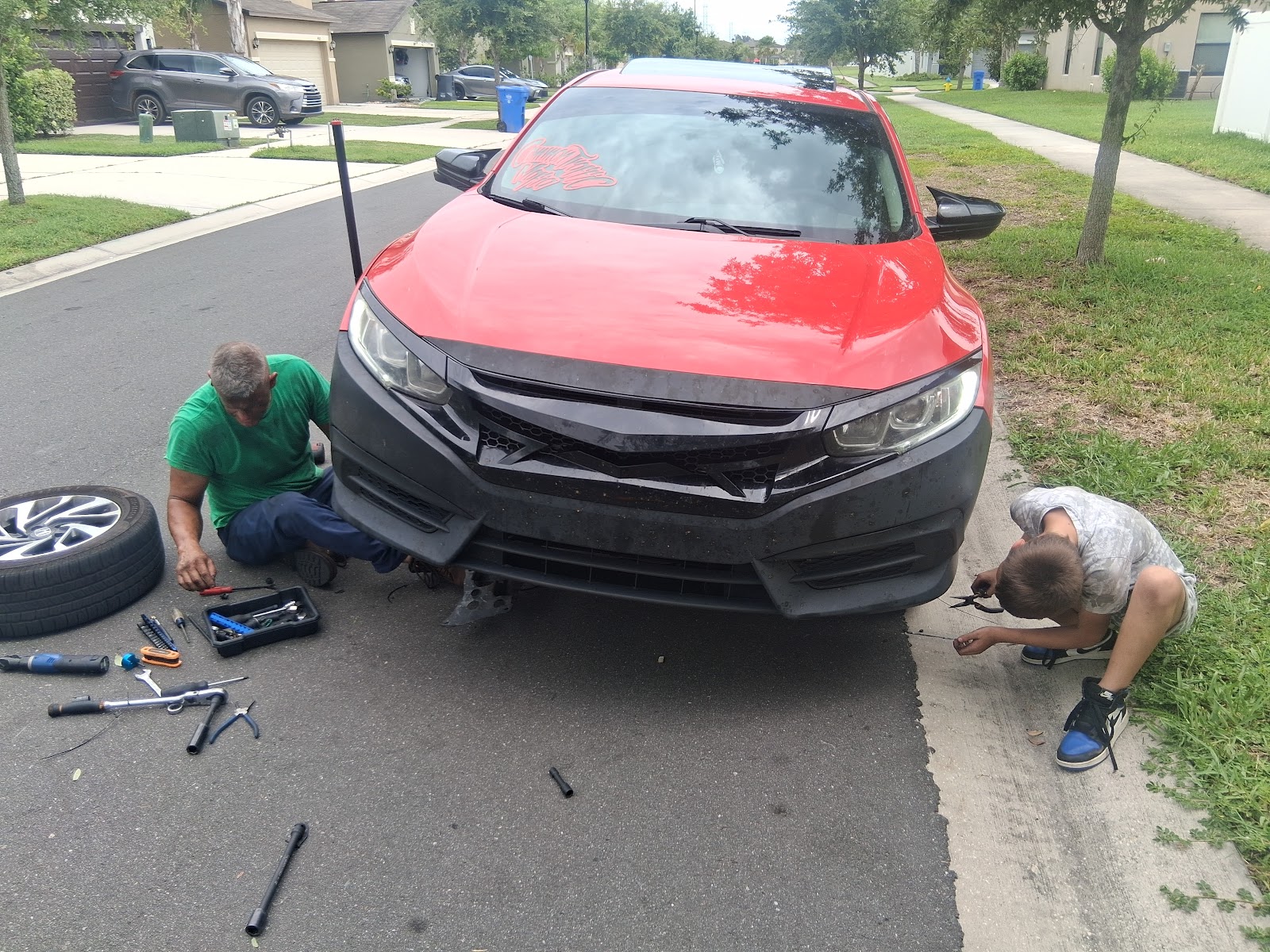 Two mechanics working on a red Honda Civic in a residential driveway